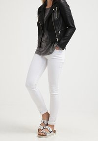 Giacca da moto nera in pelle con dettagli zip, t-shirt grigia, jeans skinny bianchi e sandali metallici con fasce e suola piatta.
