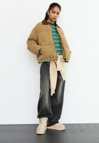 Ung kvinde iført en brun dunjakke, grønstribet skjorte, løse sorte jeans, beige støvler og en lys trøje bundet om livet.