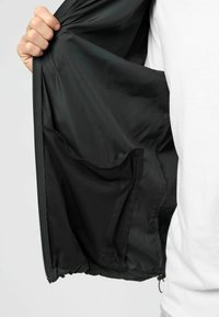 Inuti den svarta jackan finns ett mjukt foder och en meshnätficka. Dragkedjan är synlig längs kanten, med elastisk nederkant.