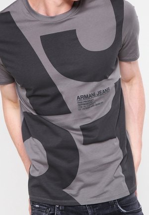 Šedé tričko s krátkým rukávem, na němž jsou velké abstraktní černé tvary. Text na tričku uvádí "Armani Jeans" a pokyny k údržbě.
