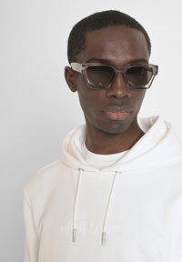Gafas de sol grises con un marco geométrico, con brazos transparentes, llevadas sobre un sudadera blanca con capucha que presenta letras en relieve. Textura suave.