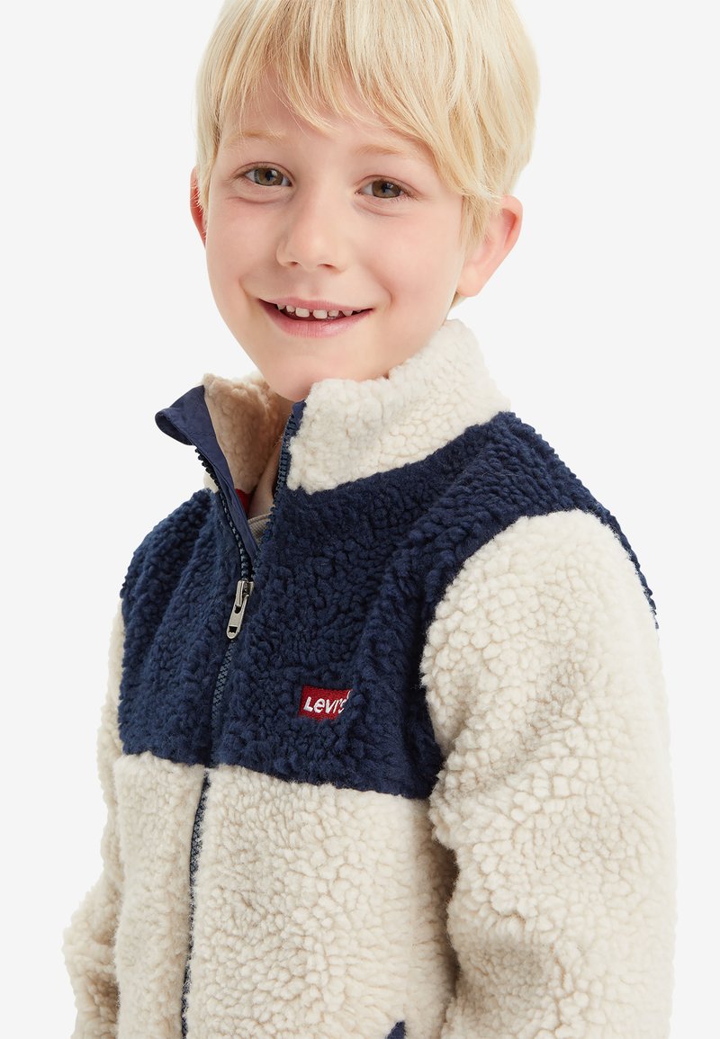 Chaqueta de forro polar con sección superior en azul marino, sección inferior en crema, cremallera frontal completa, cuello alto y un pequeño logo rojo.