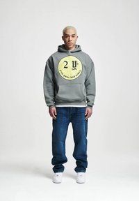 Grå hettegenser med et sirkulært gult grafikk, mørkeblå jeans og hvite joggesko. Hettegenseren har en frontlomme og en avslappet passform.
