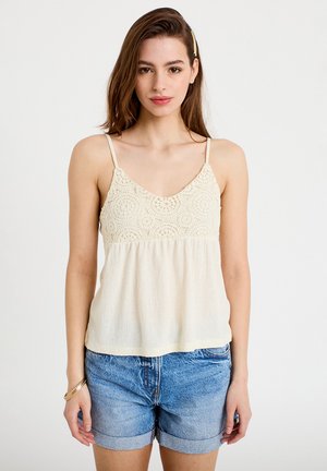 Kvinde iført en cremefarvet hæklet ærmeløs top og oppustede denimshorts, med langt brunt hår og en guldfarvet hårnål.