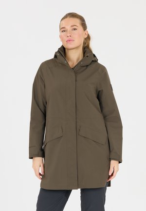 Femme portant un manteau à capuche olive vert longueur genou avec poches avant et fermeture éclair, debout devant un fond blanc uni.