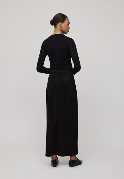 Schwarzer Wildleder-Maxi-Rock mit einem Rückennaht-Detail, kombiniert mit einem schwarzen Langarmoberteil. Modell mit dem Rücken zur Kamera, trägt schwarze flache Schuhe.