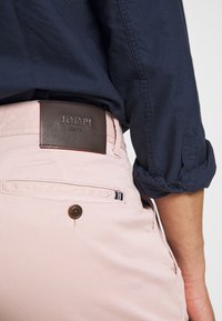 Jasnoróżowe spodnie z brązowym skórzanym paskiem z wytłoczonym logo, bocznymi kieszeniami i pojedynczym zapięciem na guzik. Widoczny rękaw granatowej koszuli.