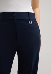 Marineblaue Hosen mit glatter Textur, ausgestattet mit einem Knopfverschluss und einem silbernen rechteckigen Akzent in der Nähe des Taillenbunds.