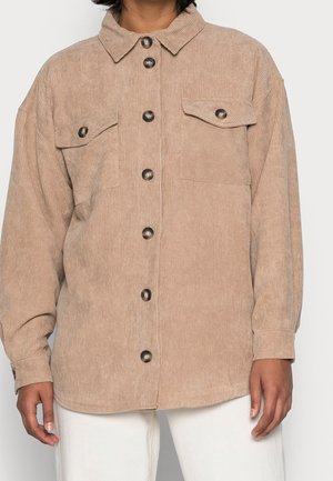 Chemise beige en velours côtelé boutonnée avec deux poches poitrine à rabat, portée sur un pantalon blanc, montrée du cou aux hanches.