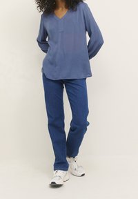 Blaues, langärmliges Oberteil mit V-Ausschnitt und subtiler Textur, kombiniert mit gerade geschnittenen blauen Jeans und weißen Sneakern mit schwarzen Akzenten.