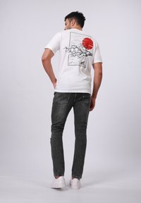 Camiseta blanca con un gráfico negro de olas y un sol rojo en la parte posterior, combinada con jeans gris oscuro y zapatillas blancas. Diseño simple.