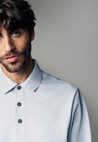Cămașă polo de culoare albastru deschis, dintr-un material texturat, cu trei nasturi închiși la culoare și un guler clasic. Prim-plan al cămășii purtate de un model.
