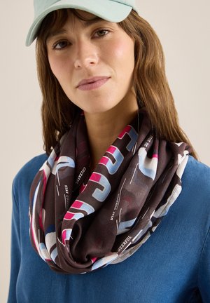 Žena nosí světle modrou čepici, modrý top a barevnou nekonečnou šálu s nápisy a geometrickými vzory kolem krku.