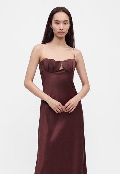 Jeune femme aux longs cheveux foncés porte une robe sans manches brun foncé avec de fines bretelles et un décolleté festonné sur un fond clair uni.