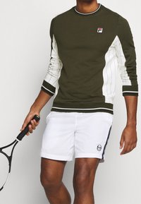 Sudadera verde oliva con paneles blancos y puños a rayas, combinada con pantalones cortos blancos que presentan un logo y acentos en navy. Sosteniendo una raqueta de tenis.