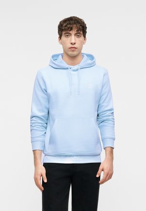 Joven de pie, vestido con una sudadera azul claro con bolsillo frontal y pantalones negros, contra un fondo gris liso.