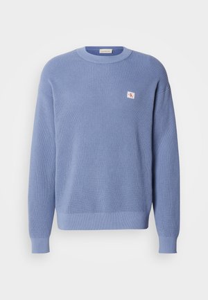 Gaiši zils adīts džemperis ar ribotu tekstūru, apaļu kakla izgriezumu un nedaudz lielāku piegriezumu. Uz krūtīm ir neliela logo uzlīme.