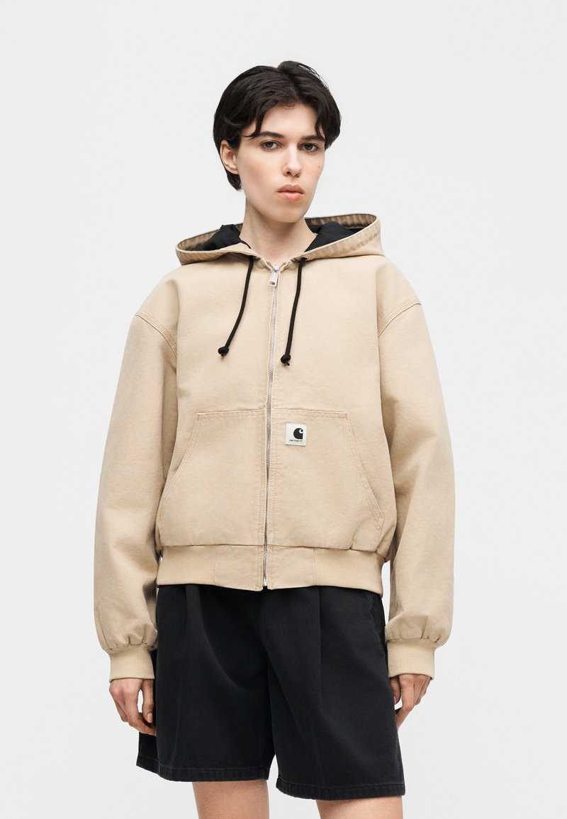 Personne portant un sweat à capuche beige zippé avec des cordons noirs et des poches avant, associé à un short noir ample sur un fond uni.