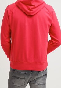 Sweat à capuche en polaire rouge avec capuche à cordon, manches raglan et poche kangourou. Associé à un jean gris avec poches arrière.
