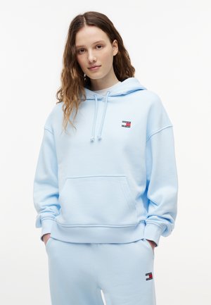 Junge Frau trägt hellblauen Hoodie und passende Jogginghose mit kleinen Tommy Jeans-Logos, Hände in den Taschen, neutraler Hintergrund.