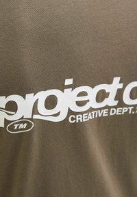 Olivgrön bomulls-t-shirt med fet vit grafisk text "project" och "CREATIVE DEPT." med ett varumärkessymbol. Slät textur.