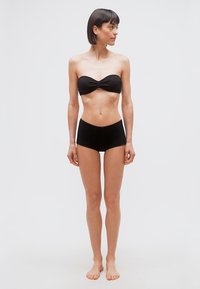 Γυναίκα που στέκεται ξυπόλυτη μπροστά σε απλό φόντο, φοράει μαύρο τοπ bandeau και αντίστοιχα σορτς, κοιτάζοντας προς τα δεξιά της.