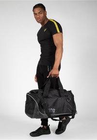 Zwarte duffelbag met meshaccenten, ritssluiting en dubbele handvaten. De tas heeft een logo en wordt gecombineerd met zwarte sportkleding en schoenen.