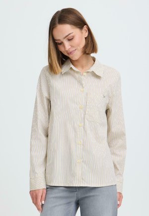 Mujer con cabello liso y castaño, vistiendo una camisa de botones a rayas beige y blanca y jeans azul claro, mirando hacia abajo con una leve sonrisa.