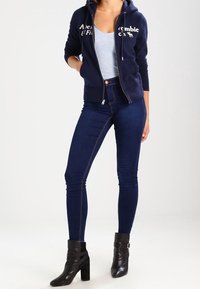 Marineblå hættejakke med lynlås og hvid tekst, lys grå tanktop, mørkeblå skinny jeans og sorte ankelstøvler med remme. Glat stof hele vejen igennem.