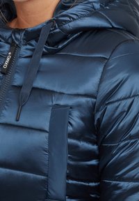 Chaqueta acolchada azul marino con textura brillante, cierre mediante cremallera, capucha ajustable y diseño acolchado horizontal con un bolsillo en el pecho.