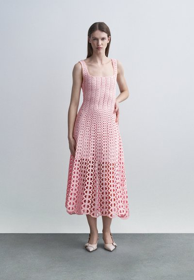 Une femme porte une robe midi en dentelle rose sans manches avec des motifs circulaires et des escarpins pointus rose pâle, posant devant un fond gris uni.