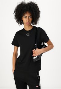 Black cotton t-shirt with short sleeves, featuring a metal logo on the chest, paired with black pants and a shiny black crossbody bag.