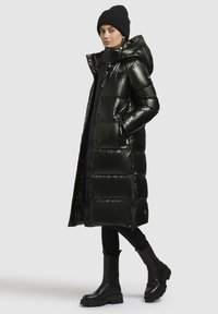 Lång svart puffjacka med glänsande yta, huva och sidofickor. Bärs över svart topp och leggings, kombinerat med svarta kängor med tjock sula.