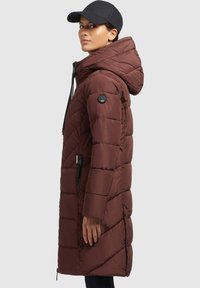 Ruskea pehmeä parka-takki, jossa on korkea kaulus, säädettävä huppu ja vetoketjulliset sivutaskut. Takissa on rakenteellinen tikkikuviointi ja sivusaumat.