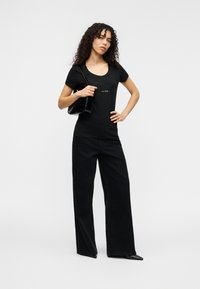 Μαύρο εφαρμοστό μπλουζάκι με στρογγυλή λαιμόκοψη και ανάγλυφο λογότυπο, συνδυασμένο με φαρδιά μαύρη ψηλόμεση παντελόνα και μικρή μαύρη τσάντα χειρός.