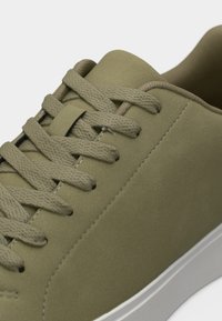 Κοντινό πλάνο αθλητικού παπουτσιού ελιάς πράσινου χρώματος με υφασμένα κορδόνια, λείο ανώτερο υλικό και λευκή λαστιχένια σόλα.