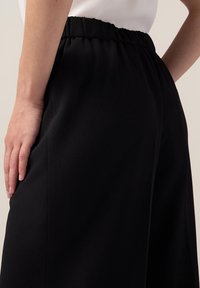 Pantalons noirs avec une taille élastique, texture lisse et poches latérales. Présentés de côté, avec une main posée sur la hanche.