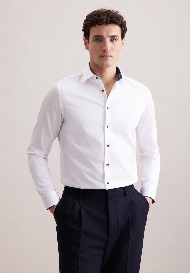 Camicia bianca a maniche lunghe con colletto blu a contrasto. Presenta bottoni scuri lungo la parte anteriore e una vestibilità su misura. Indossata con pantaloni blu.