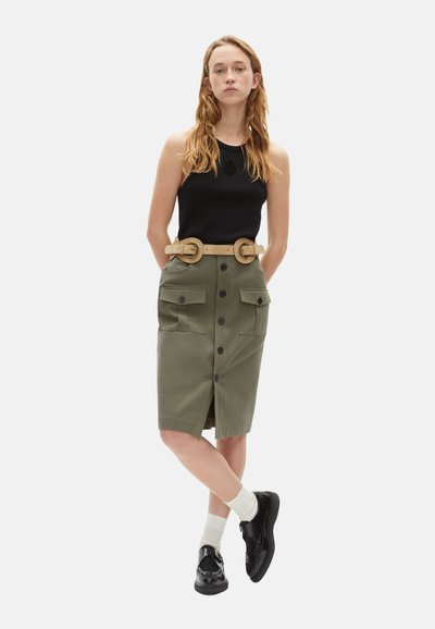 Zwart tanktop gecombineerd met een olijfgroen knielang rokje met grote zakken, knoopdetails en een brede beige riem met een dubbele gesp.