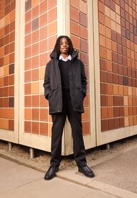 Sort parka med hætte, lagdelt over en sort sweater og hvid skjorte, kombineret med sorte bukser og sko, foran en tekstureret orange væg.