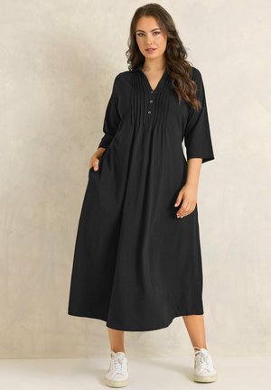 Kobieta ubrana w czarną, długą, luźną sukienkę z rękawami do połowy przedramienia oraz białe sportowe buty, stojąca na tle jasnego, jednolitego tła.