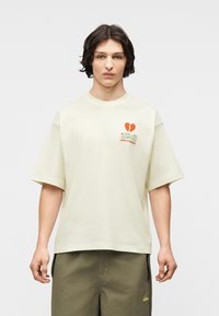 Νεαρός με σκοτεινά μαλλιά, που φοράει εκρού κοντομάνικο πουκάμισο με σπασμένη καρδιά και κείμενο στο στήθος και ελαιδί παντελόνι με λογότυπο.