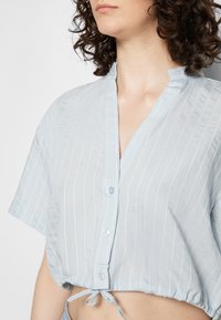 Camisa curta azul clara com mangas curtas, listas brancas verticais, frente abotoada e detalhe com cordão na bainha.