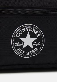 Sac en tissu noir avec un logo circulaire blanc comportant une étoile et le texte "Converse Chuck Taylor All Star". Fermeture éclair visible.