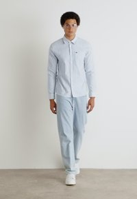 Hellblau gestreiftes Button-Up-Hemd mit langen Ärmeln, kombiniert mit hellblauen Hosen. Model trägt weiße Sneaker und runde Brillen.