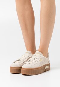 Beige mockaskplatformsskor med en texturerad krämfärgad sula, som har platta snören och en subtil guldlogo detaljerad på tungan.