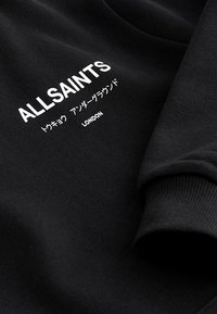 Sort stof med hvid tekst, der siger ALLSAINTS, japanske tegn og LONDON, der viser en del af en ærme med ribstrikket manchet.
