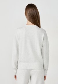 Woman with straight brown hair wearing a light gray sweater and matching pants, standing with her back facing the camera on a white background.