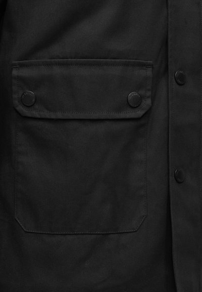 Μαύρο μπουφάν από ύφασμα με μεγάλο τσέπη στο στήθος, κλεισμένη με δύο μαύρα κουμπιά τύπου κλικ και ματ υφή. Απλός, λειτουργικός σχεδιασμός.
