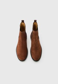 Par de botas Chelsea de ante marrón con paneles laterales de elastó negro y tiradores, exhibidas sobre un fondo gris claro.
